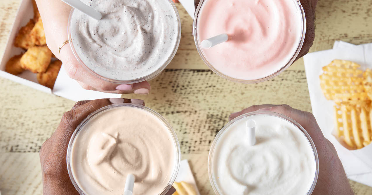 Top-view of four hands holding milkshakes in different flavors, with waffle fries and chicken nuggets in the background.