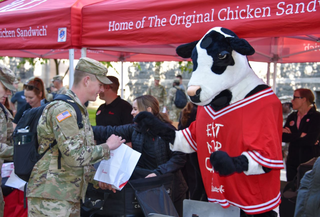 Chick-fil-A cow mascot greets a U.S. soldier at an outdoor event under a red tent labeled 'Home of The Original Chicken Sandwich.'