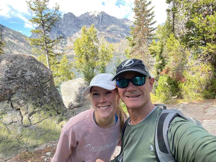 Father and daughter, Glen and Olivia Efford, smile while hiking in the mountains, surrounded by trees and rocky terrain with peaks in the background.
