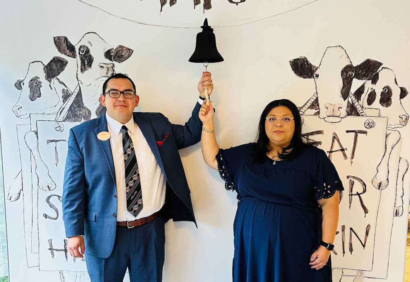 A man in a blue suit and a woman in a blue dress hold a rope attached to a bell, standing in front of cow drawings on a wall.