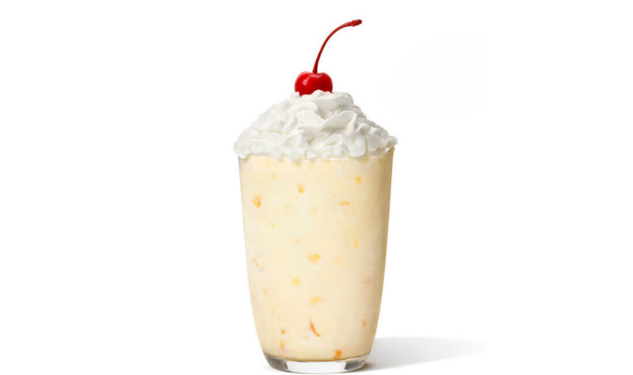 Peach milkshake in a glass topped with whipped cream and a cherry.