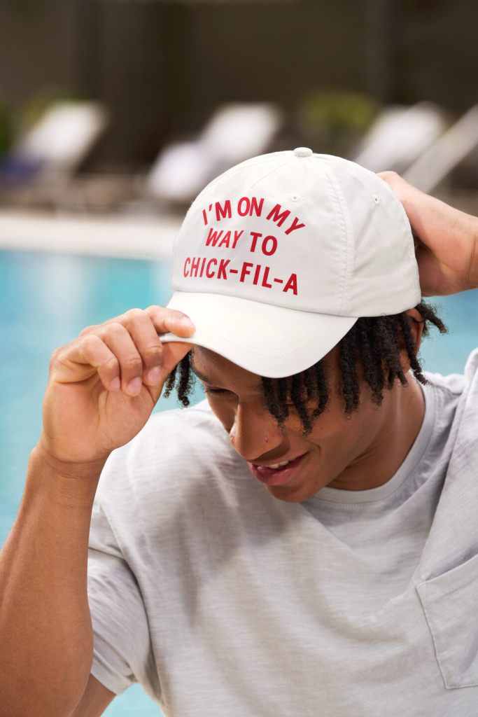 Man smiles while adjusting a white baseball cap with red text that reads “I’M ON MY WAY TO CHICK-FIL-A,” near a swimming pool.