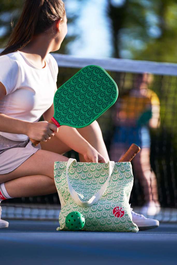 Person crouched on a court holding a green pickleball paddle with a patterned tote bag and ball nearby, another player in the background.