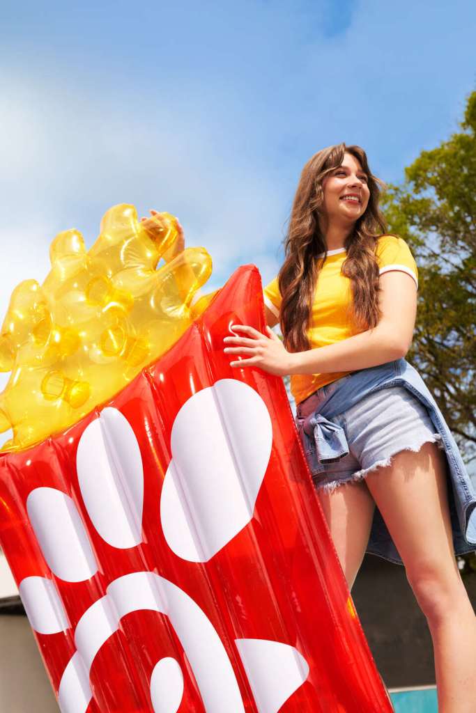 Smiling woman in a yellow shirt holds a large inflatable pool float shaped like Chick-fil-A waffle fries, outdoors under a blue sky.
