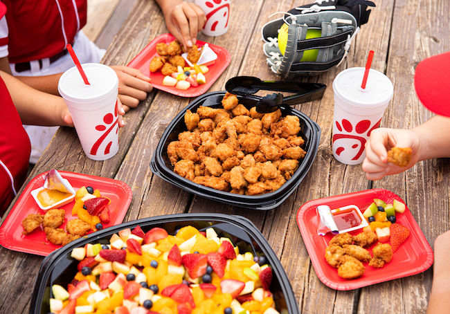 Baseball players sitting at a table with trays of chicken nuggets and fruit, alongside drinks and a baseball glove.

