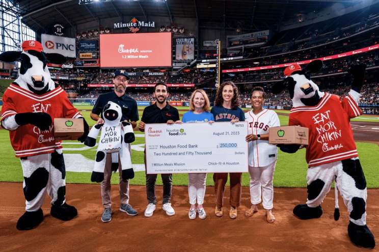 Group of people at Minute Maid Park hold a $250,000 check from Chick-fil-A Houston to Houston Food Bank, flanked by two Chick-fil-A cow mascots in red jerseys.
