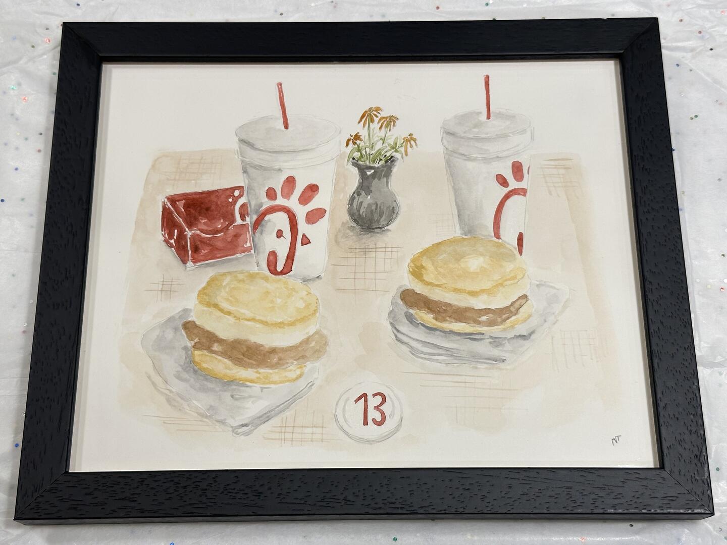 Framed watercolor painting of a Chick-fil-A meal with two chicken biscuits, two drinks with straws, a red box, a small vase of flowers, and a table marker with the number 13.