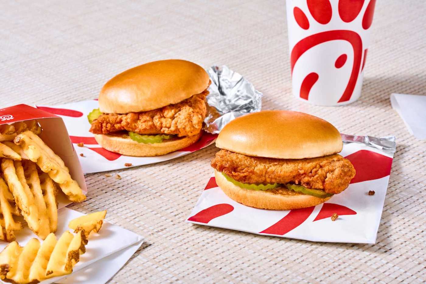 Two Chick-fil-A® chicken sandwiches with waffle fries and a drink on a light woven surface.