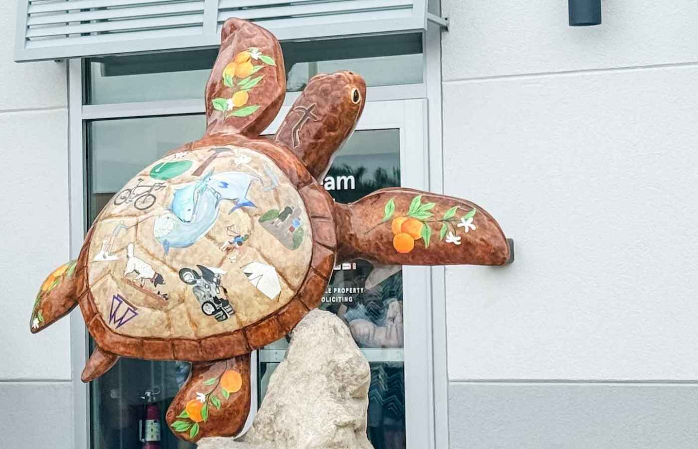 Colorful sea turtle sculpture with painted images and orange blossoms on its shell, mounted on a rock outside a building.