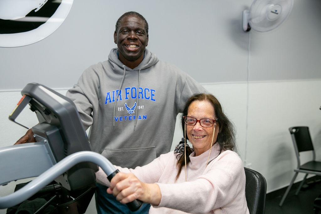 A man in an Air Force Veteran hoodie stands next to a woman seated on exercise equipment.

