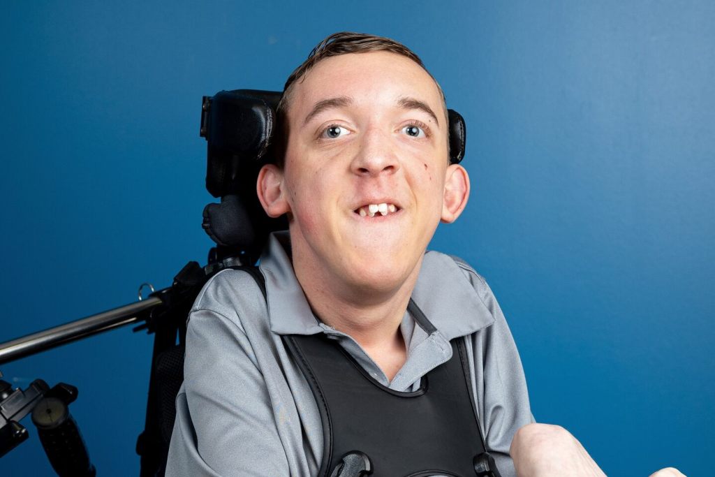 A man in a wheelchair smiles at the camera in front of a blue background.