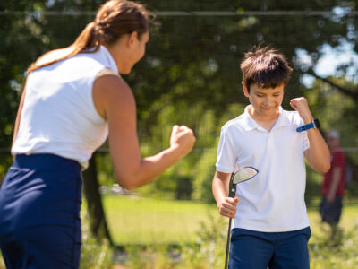 A boy celebrates with a golf club as a woman cheers in a sunny outdoor setting.