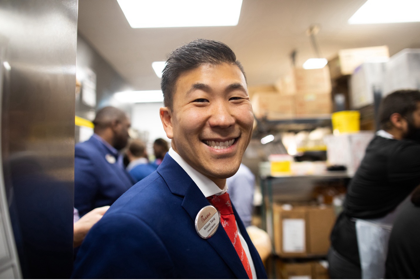 Operator Wilson Yang smiling in a Chick-fil-A kitchen, wearing a suit and name badge, surrounded by team members.