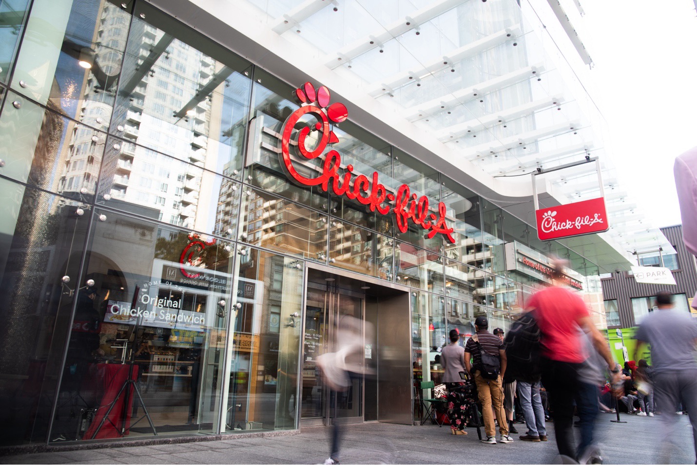 Customers lining up outside a glass-front Chick-fil-A restaurant in Canada, with city buildings reflected in the windows.