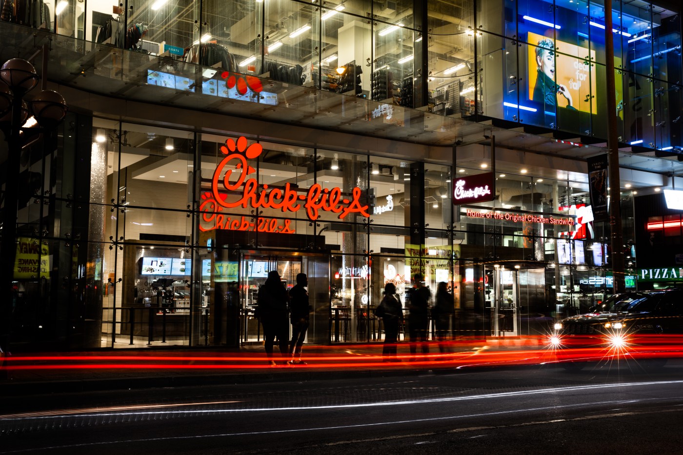 Exterior of a Chick-fil-A restaurant at night in downtown Toronto with glowing signage and light trails from passing cars.
