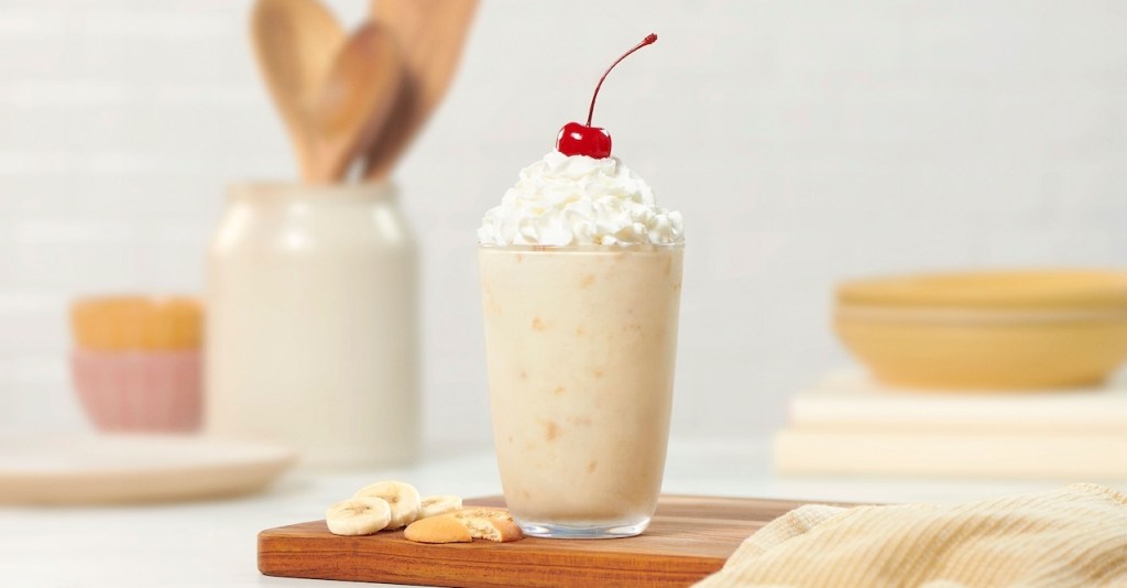 A banana pudding milkshake with whipped cream and a cherry on top, surrounded by banana slices and cookies.