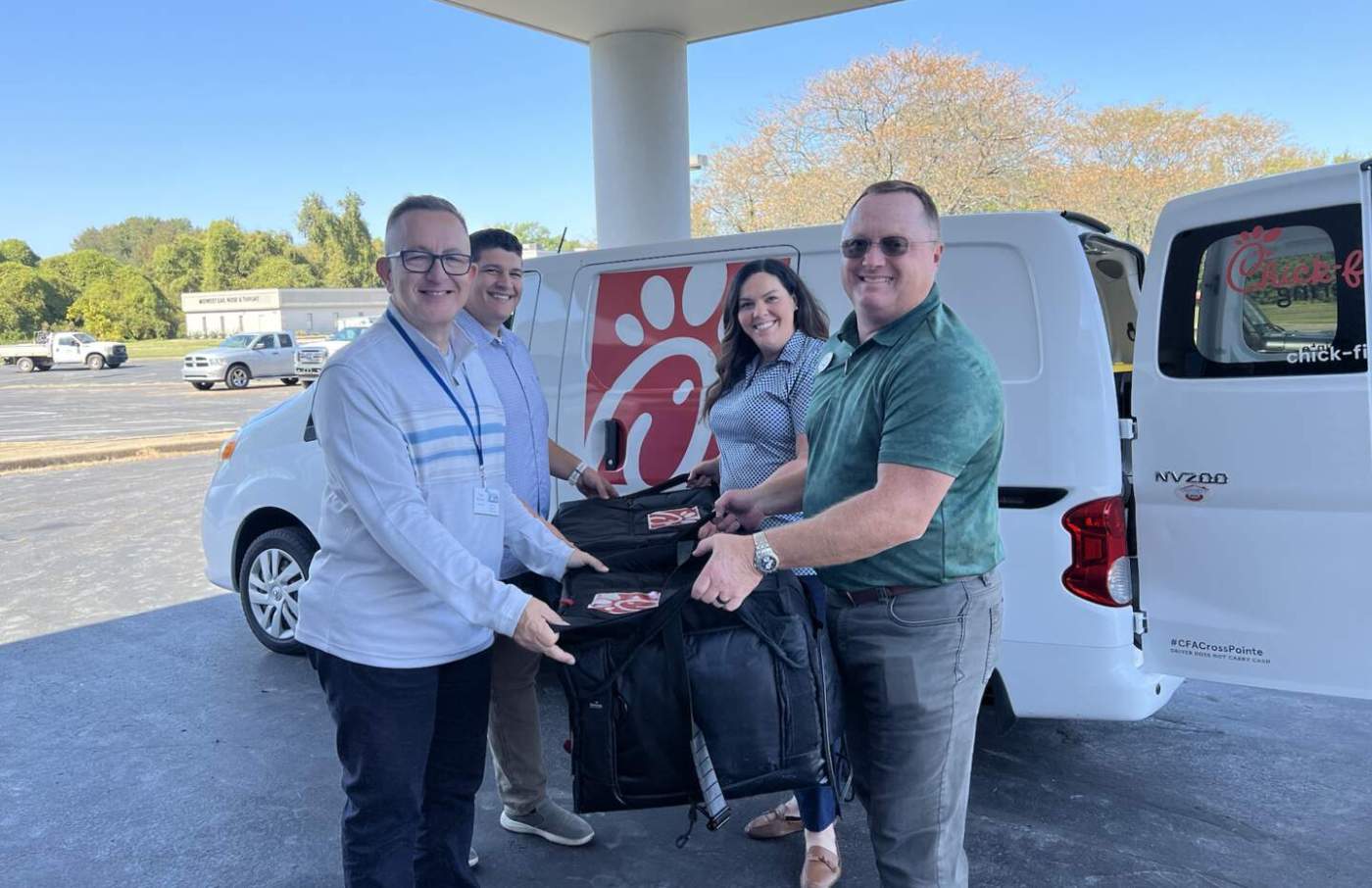 Four people stand in front of a white van with a Chick-fil-A logo, holding insulated bags.