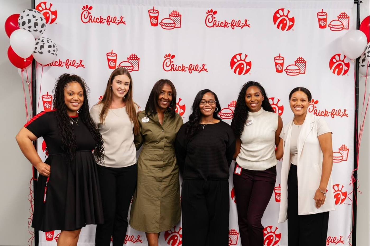 Six women pose smiling in front of a Chick-fil-A branded step-and-repeat banner with red and white balloons on either side.