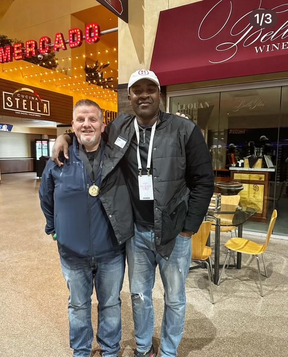 Two men stand arm-in-arm smiling at an indoor venue with signs for restaurants and wine bar in the background.