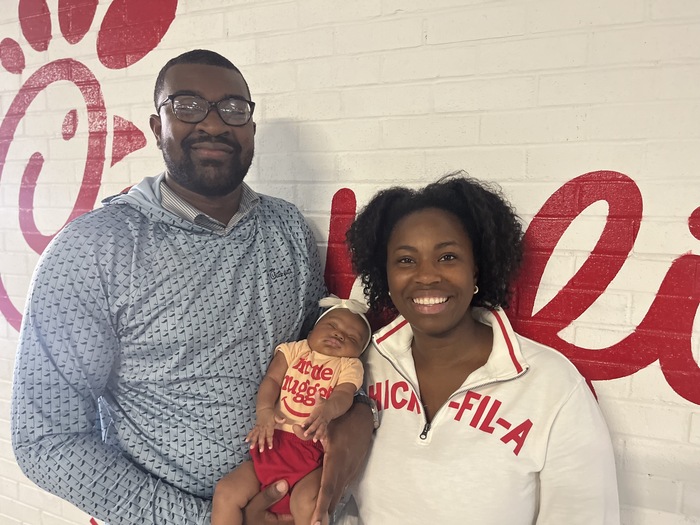 Man and woman smile while holding a sleeping baby in front of a Chick-fil-A wall mural.


