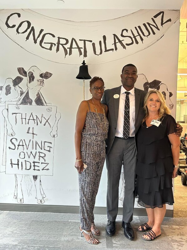 Man in a suit stands with two women in front of a Chick-fil-A wall mural that says 'Congratulashunz' and 'Thanx 4 Saving Our Hidez.