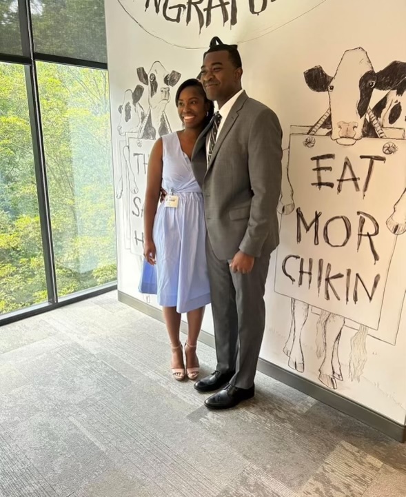 Man in a suit poses with a woman in a blue dress in front of a Chick-fil-A themed wall with 'Eat Mor Chikin' message and cartoon cows.

