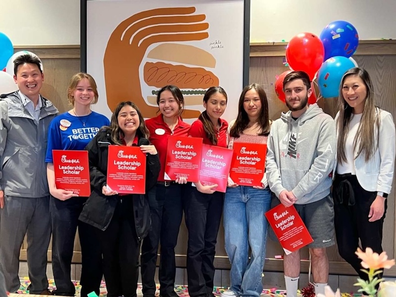 A group of Chick-fil-A Leadership Scholars and team members pose with red award plaques in front of a sandwich-themed backdrop and colorful balloons.