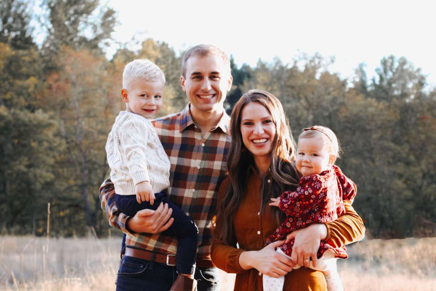 A smiling couple stands outdoors with their young son and daughter, posing in front of a wooded background on a sunny day.