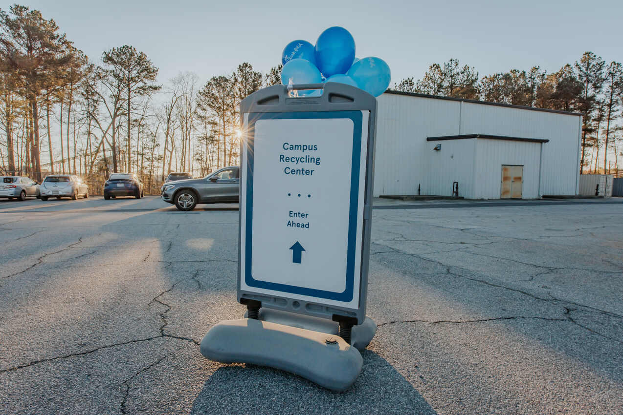 Sign in a parking lot directing to the Campus Recycling Center, with blue balloons attached.