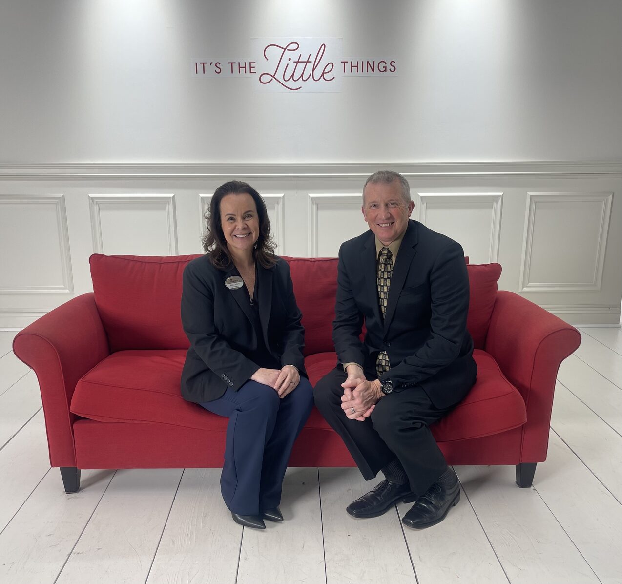 Kasi Corbett and her husband smile while sitting on a red couch beneath a wall sign that reads “It’s the Little Things.”