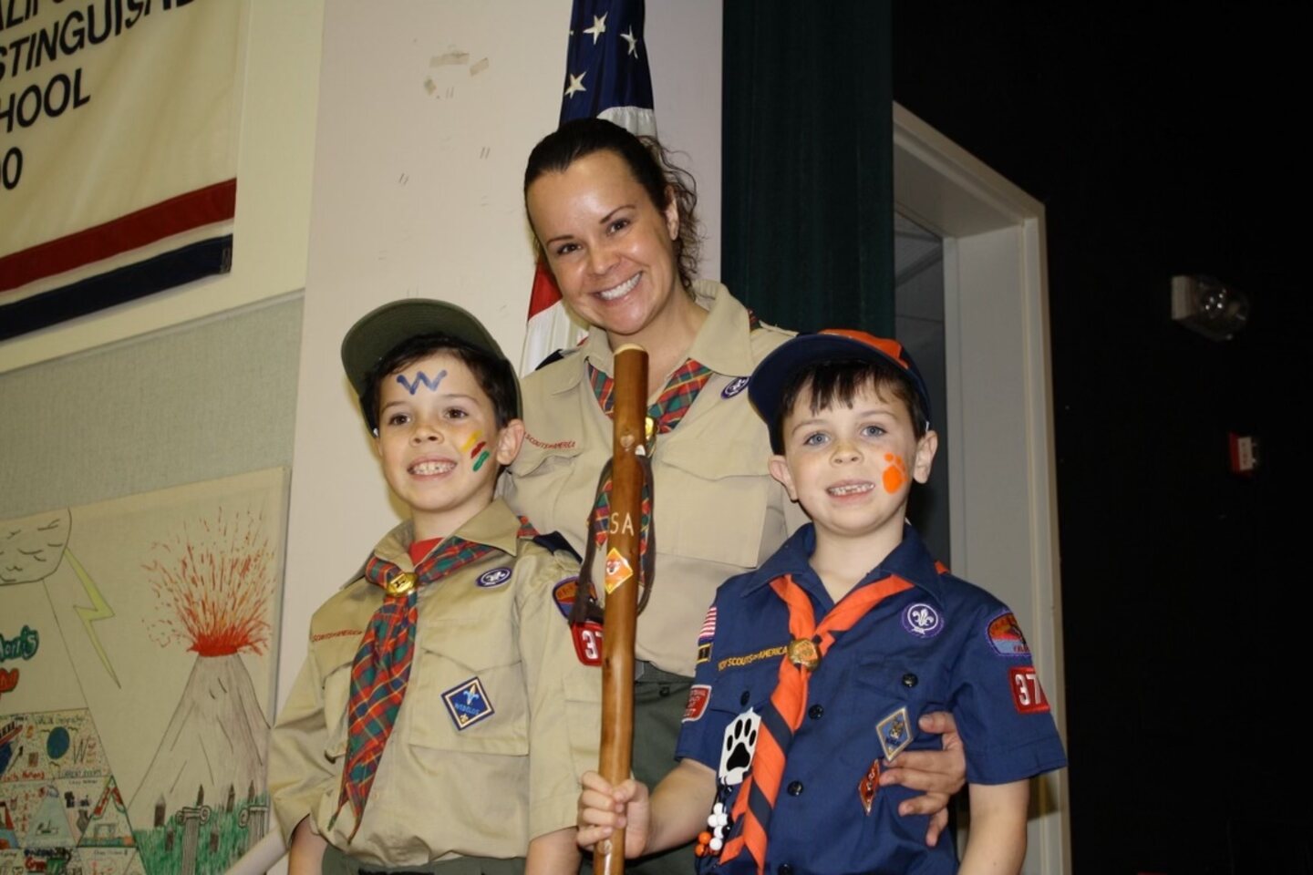 Kasi Corbett, in a scout uniform, smiles with two young boys in scout uniforms with painted faces at an indoor ceremony.