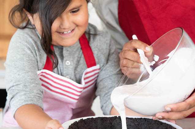 A little girl smiling helping her mother pour filling into pie crust.