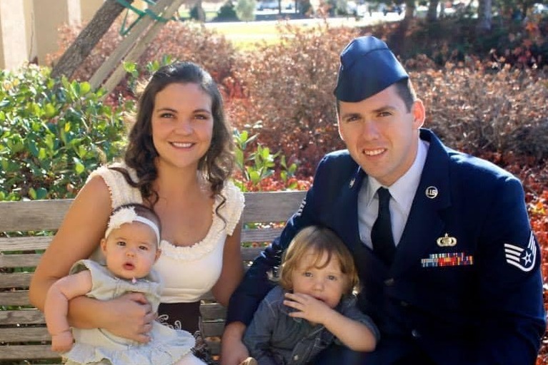 A man in a U.S. Air Force uniform sits on a bench with his wife and two young children, smiling outdoors on a sunny day.