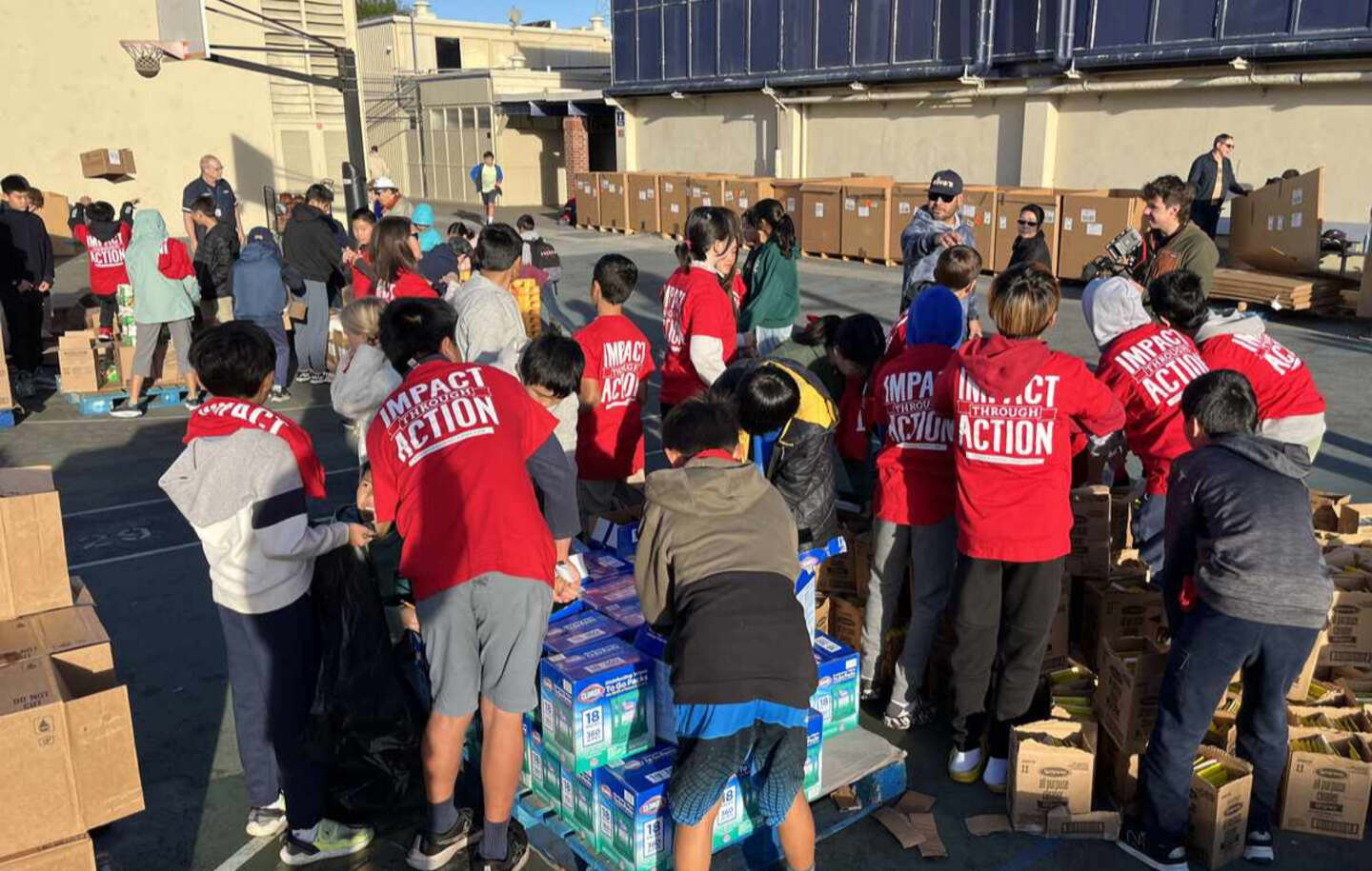 A group of people, many in red "IMPACT THROUGH ACTION" shirts, organizing and packing boxes outdoors.

