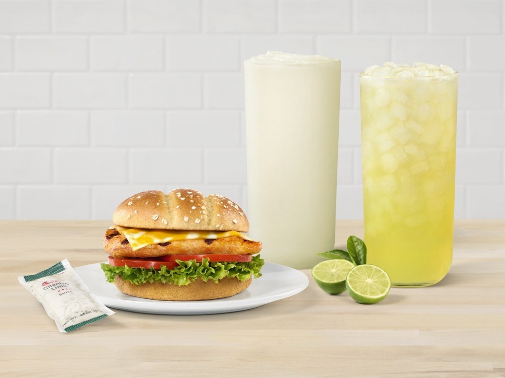 A Grilled Spicy Deluxe Sandwich on a plate with two Seasonal Key Lime Beverages, and limes on the side.