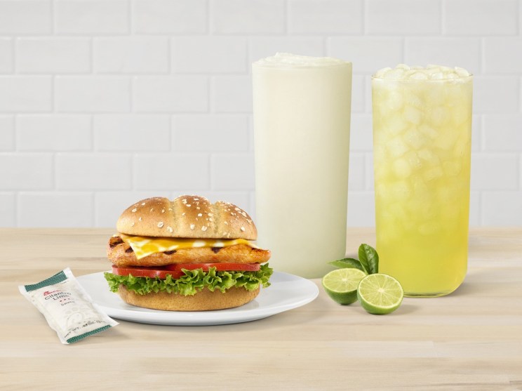 A Grilled Spicy Deluxe Sandwich on a plate with two Seasonal Key Lime Beverages, and limes on the side.