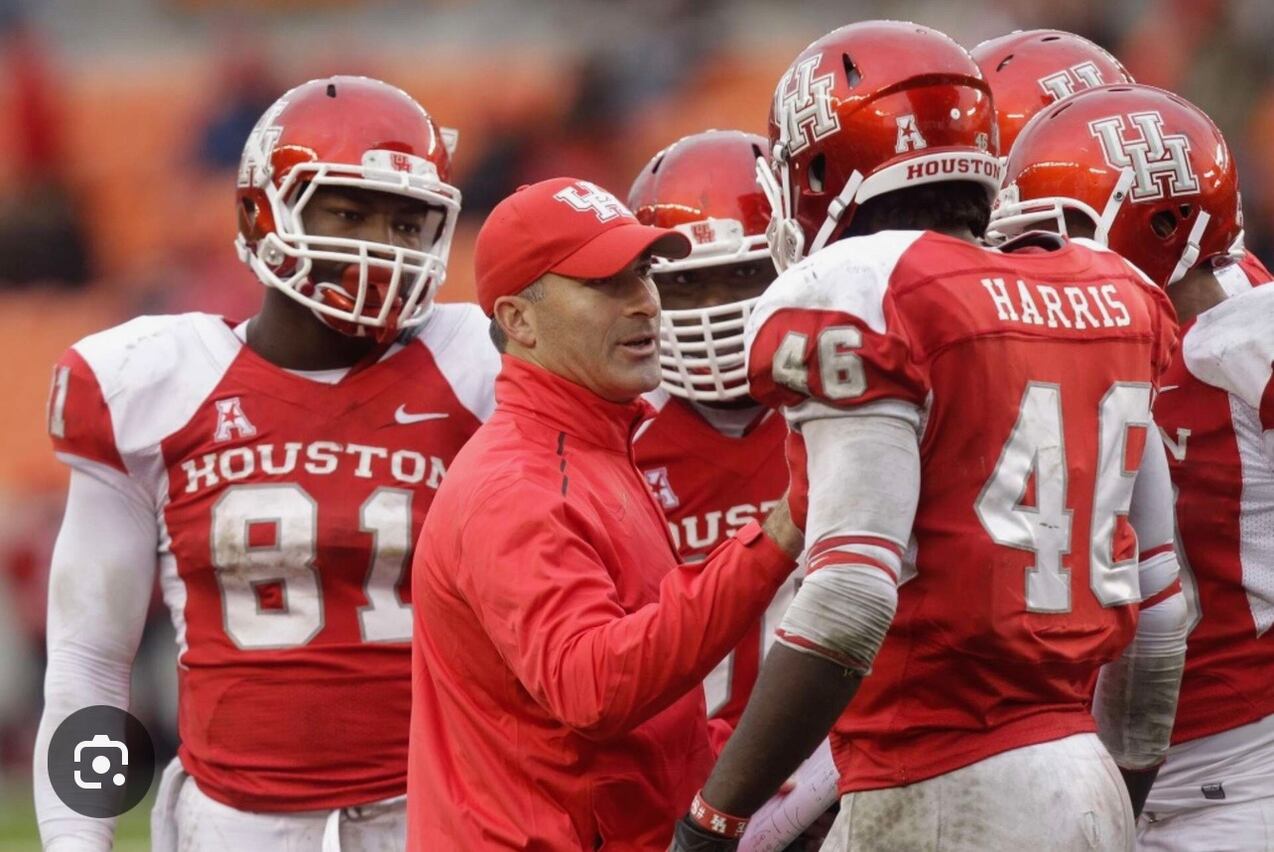 Houston football coach in red jacket talks with players in red uniforms and helmets on the sideline during a game.
