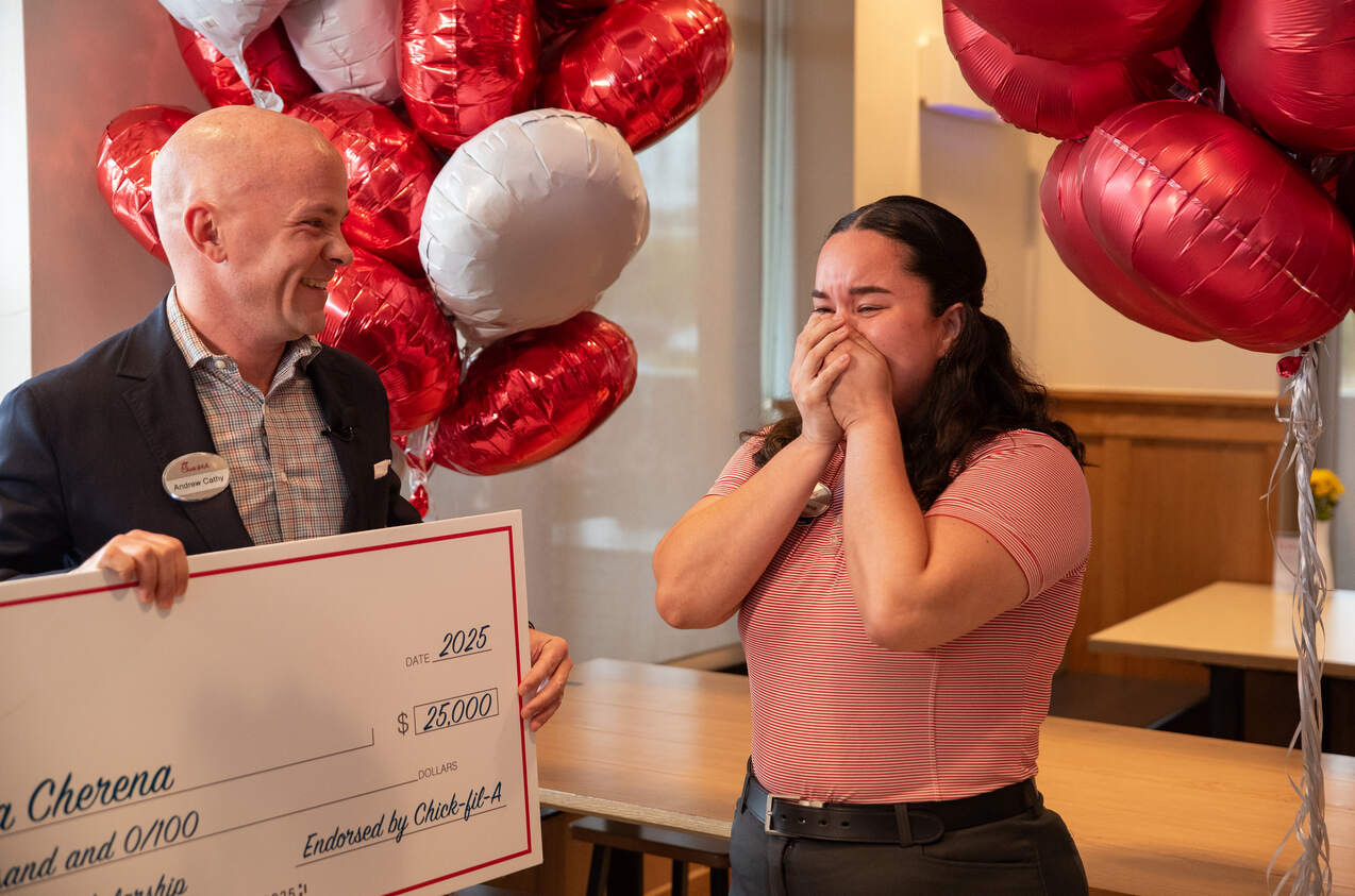 A man presents a large check to a woman, who reacts with emotion, surrounded by balloons.