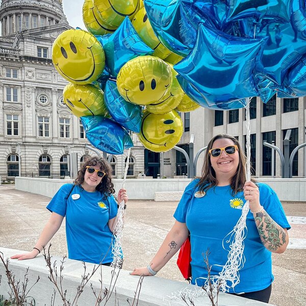 Two people in blue T-shirts holding blue star and yellow smiley face balloons in front of a classical building.