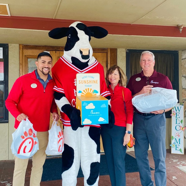 Four people, including a cow mascot in a red jersey, pose with bags and a box outside a building.