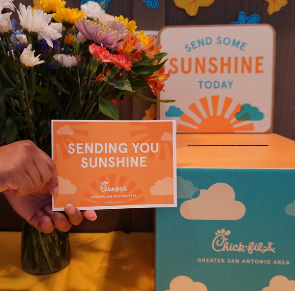 A hand holds an orange card saying "SENDING YOU SUNSHINE" near flowers and a turquoise box with clouds and the Chick-fil-A logo.