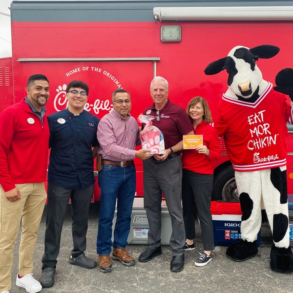 A group of people, including a person in a cow costume, stand in front of a Chick-fil-A food truck holding a gift.