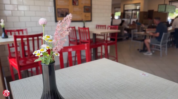 A Chick-fil-A restaurant table with a black vase holding small flowers; red chairs and a tiled wall are visible in the background.
