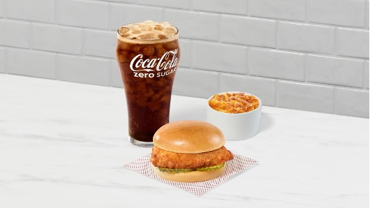 Chicken sandwich with macaroni and cheese and a glass of Coca-Cola Zero Sugar on a white surface.
