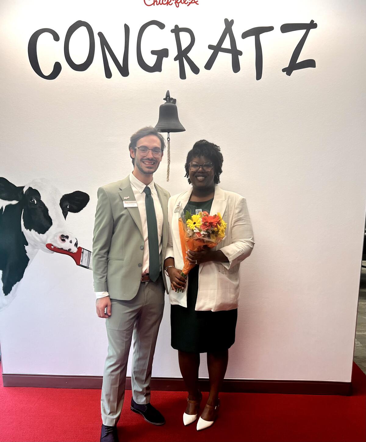 Chantay Bollinger and another person stand smiling in front of a wall with "CONGRATZ" written in black and a cow illustration.