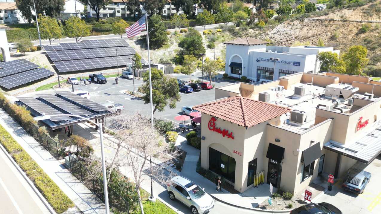 Aerial view of a Chick-fil-A restaurant with solar panels over a parking area and surrounding commercial buildings.