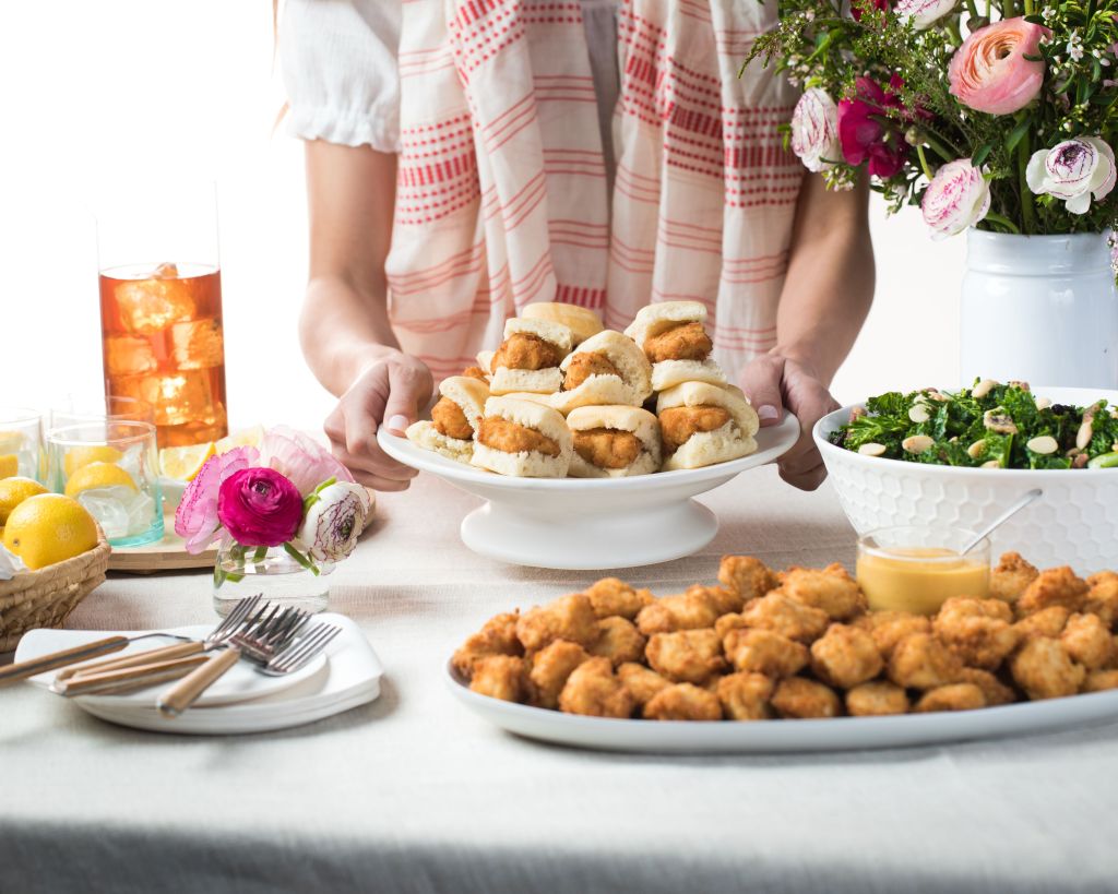 Person setting a platter of Chick-fil-A Chick-n-Minis® Tray on a table with Chick-fil-A® Nuggets Tray, a Kale Crunch Side Tray, utensils and Iced Tea