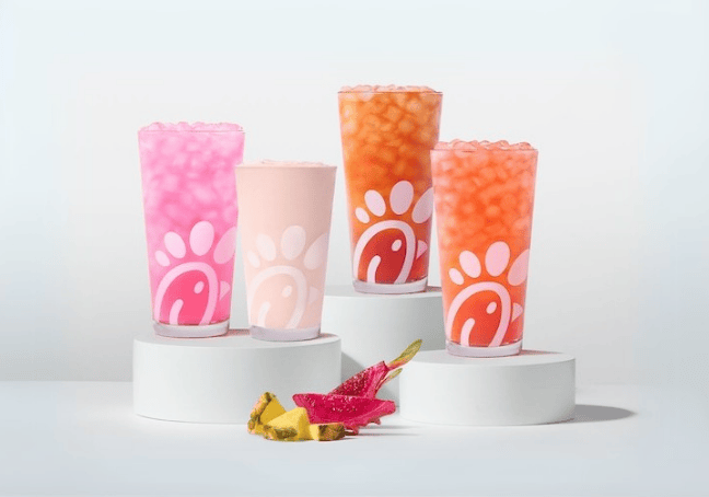 A glass of a Pineapple Dragonfruit Lemonade, Frosted Lemonade, Iced Tea and Sunjoy with pieces of pineapple and dragonfruit next to them