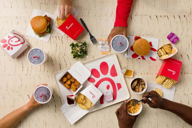 Overhead view of Chick-fil-A items, including chicken sandwiches, fries, chicken nuggets, and drinks, on a wooden table with visible hands interacting with the food.