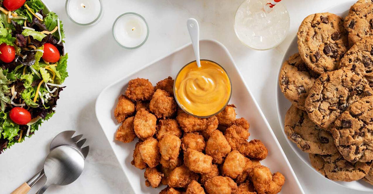 A spread of salad, chicken nuggets with dipping sauce, and chocolate chip cookies on a white tabletop.